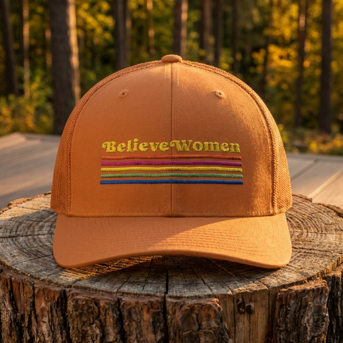 Believe Women embroidered feminist trucker hat with rainbow stripe detail, structured front, breathable mesh back, and adjustable snap closure.