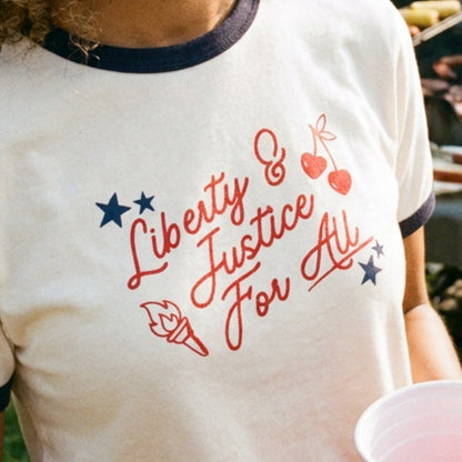 A smiling woman outdoors wearing a cream ringer T-shirt with navy trim and red script text reading 'Liberty & Justice For All,' decorated with red cherries and blue stars.