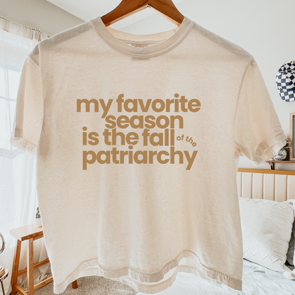 A beige short-sleeve graphic t-shirt hanging on a wooden hanger in a bedroom setting. The shirt features the text 'my favorite season is the fall of the patriarchy' printed in a warm brown sans-serif font.
