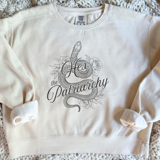 An off-white cropped sweatshirt featuring a black line art graphic of a snake wrapped around the words 'Hex the Patriarchy' with floral details, laid flat on a textured cream knit blanket.