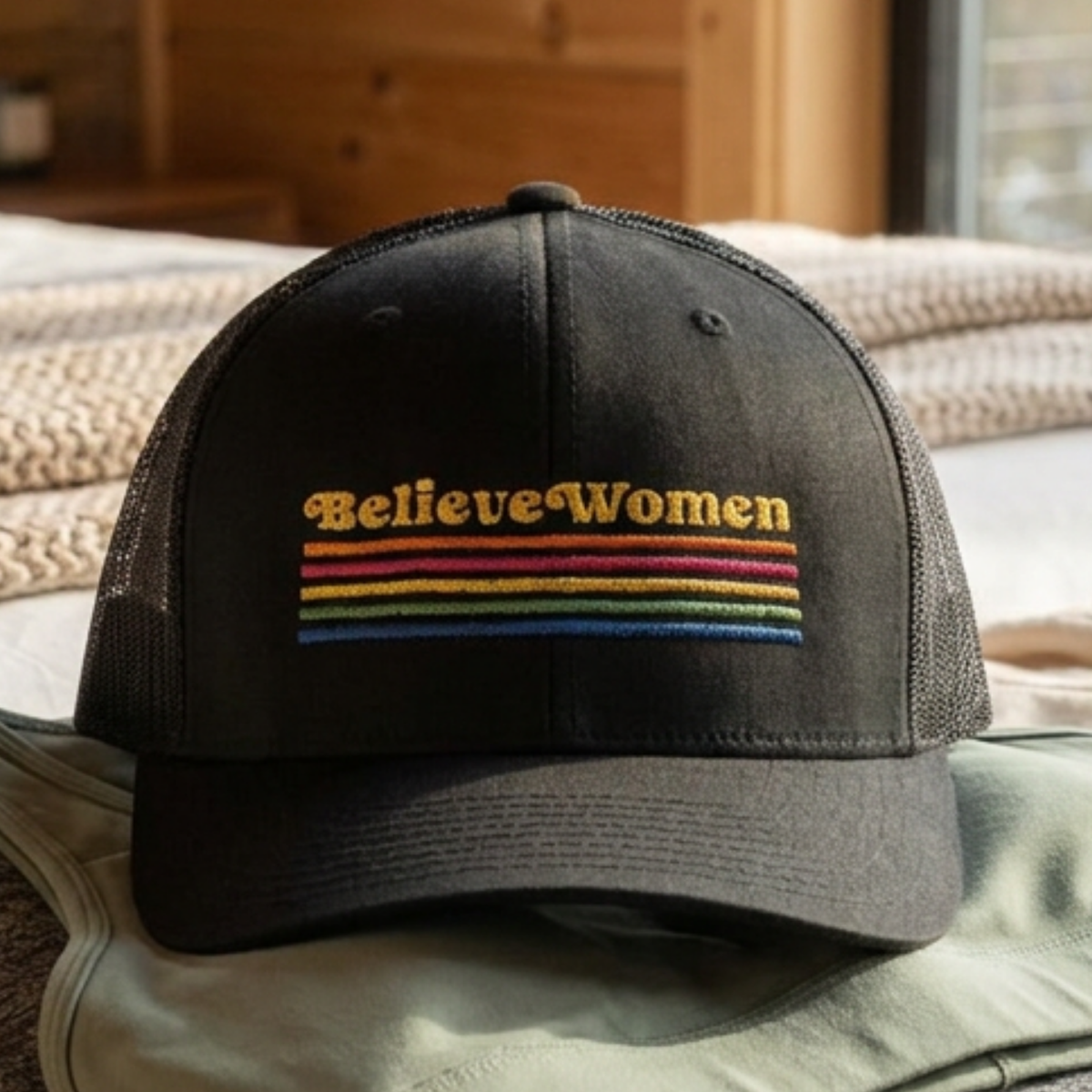 Believe Women embroidered feminist trucker hat with rainbow stripe detail, structured front, breathable mesh back, and adjustable snap closure.