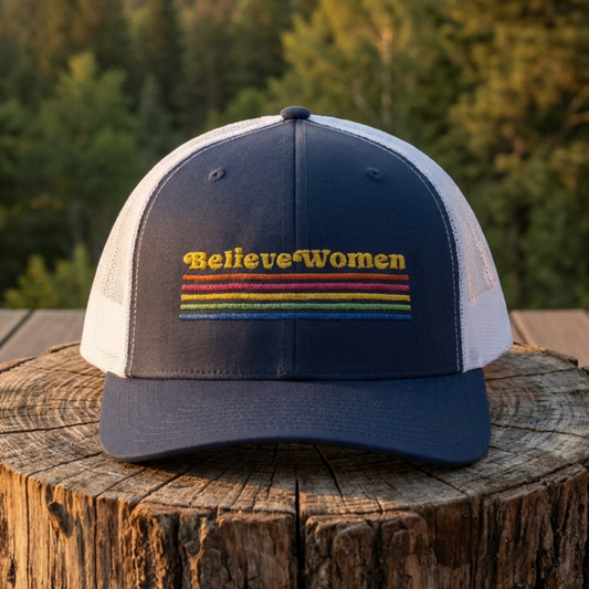 Believe Women embroidered feminist trucker hat with rainbow stripe detail, structured front, breathable mesh back, and adjustable snap closure.