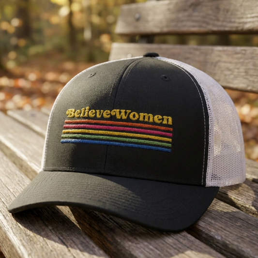 Believe Women embroidered feminist trucker hat with rainbow stripe detail, structured front, breathable mesh back, and adjustable snap closure.
