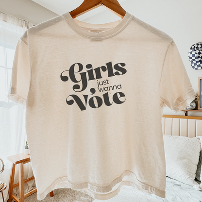 A cream-colored short-sleeve t-shirt with a black graphic reading 'Girls just wanna Vote' is held up on a wooden hanger by a hand in a bedroom setting.