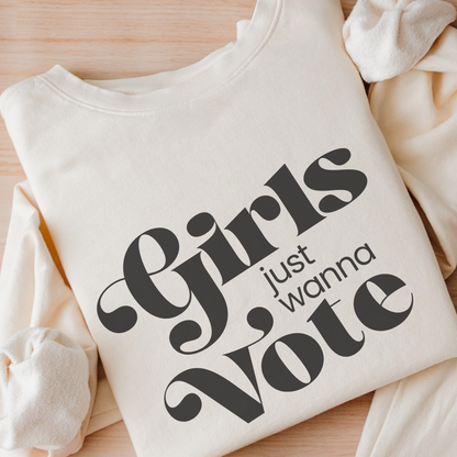 An off-white crewneck sweatshirt with the slogan 'Girls just wanna Vote' printed in black script, styled on a light wood background.