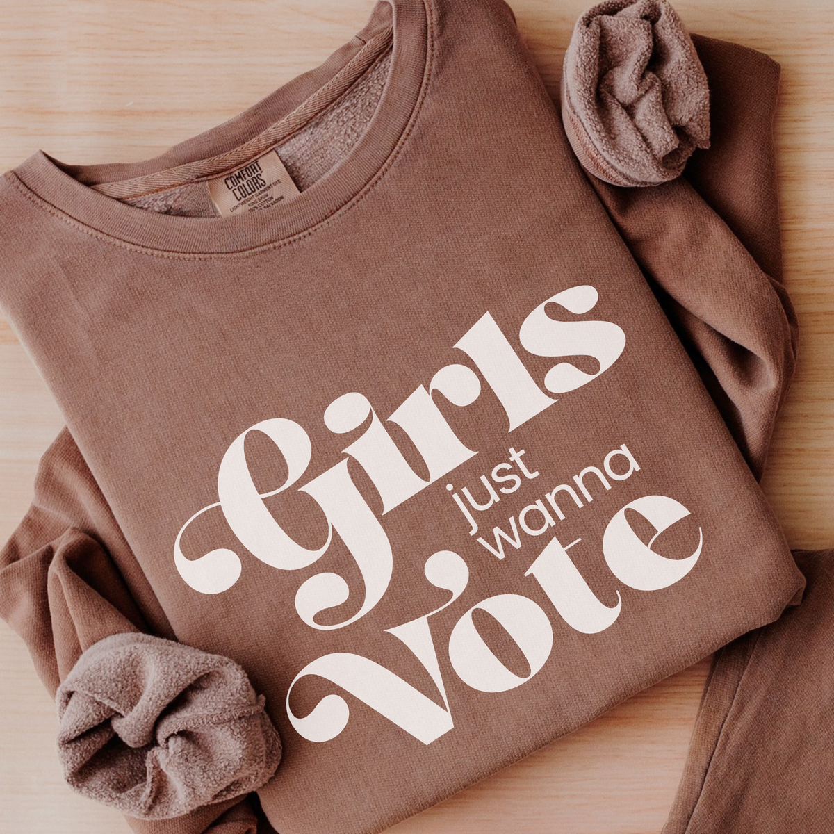 A brown crewneck sweatshirt laid flat on a light wood surface, featuring the phrase 'Girls just wanna Vote' printed in large white script across the front. 