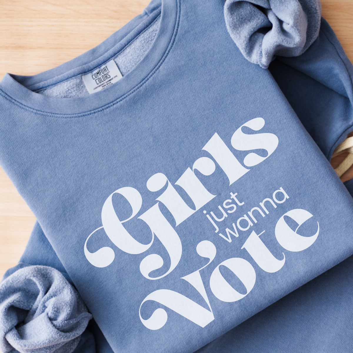 A folded dusty blue sweatshirt on a light wood background, featuring the slogan 'Girls just wanna Vote' printed in white across the front.