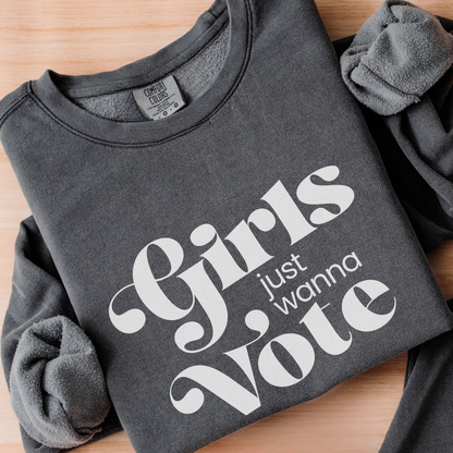 A folded dark gray fleece sweatshirt with rolled sleeves, featuring the phrase 'Girls just wanna Vote' printed in large white text on the front, laid flat on a light wood surface.