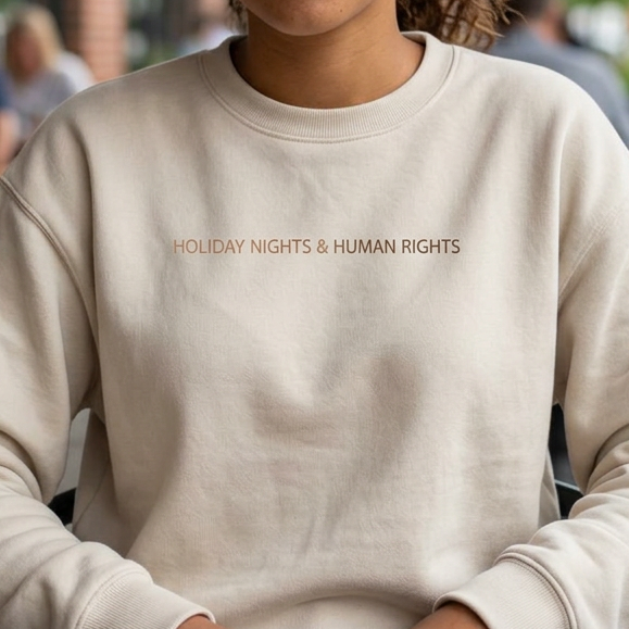 A woman sitting at a cafe table wearing a cream-colored crewneck sweatshirt with the text 'HOLIDAY NIGHTS & HUMAN RIGHTS' printed in metallic brown across the chest.