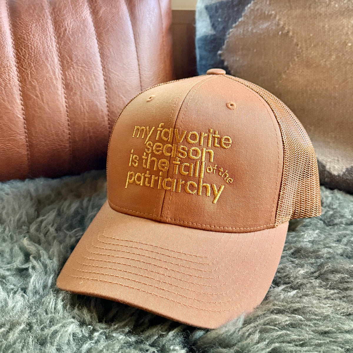 A tan trucker-style baseball cap with gold embroidered text reading 'my favorite season is the fall of the patriarchy' resting on a gray shag rug in front of a brown leather pillow.