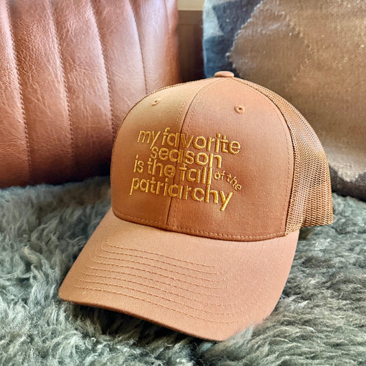 A tan trucker-style baseball cap with gold embroidered text reading 'my favorite season is the fall of the patriarchy' resting on a gray shag rug in front of a brown leather pillow.