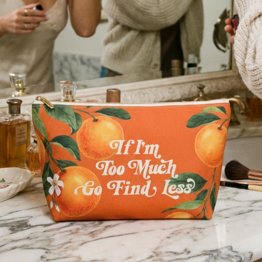 "If I'm Too Much, " Orange makeup bag with orange fruit design with white and gold zipper on a marble countertop.